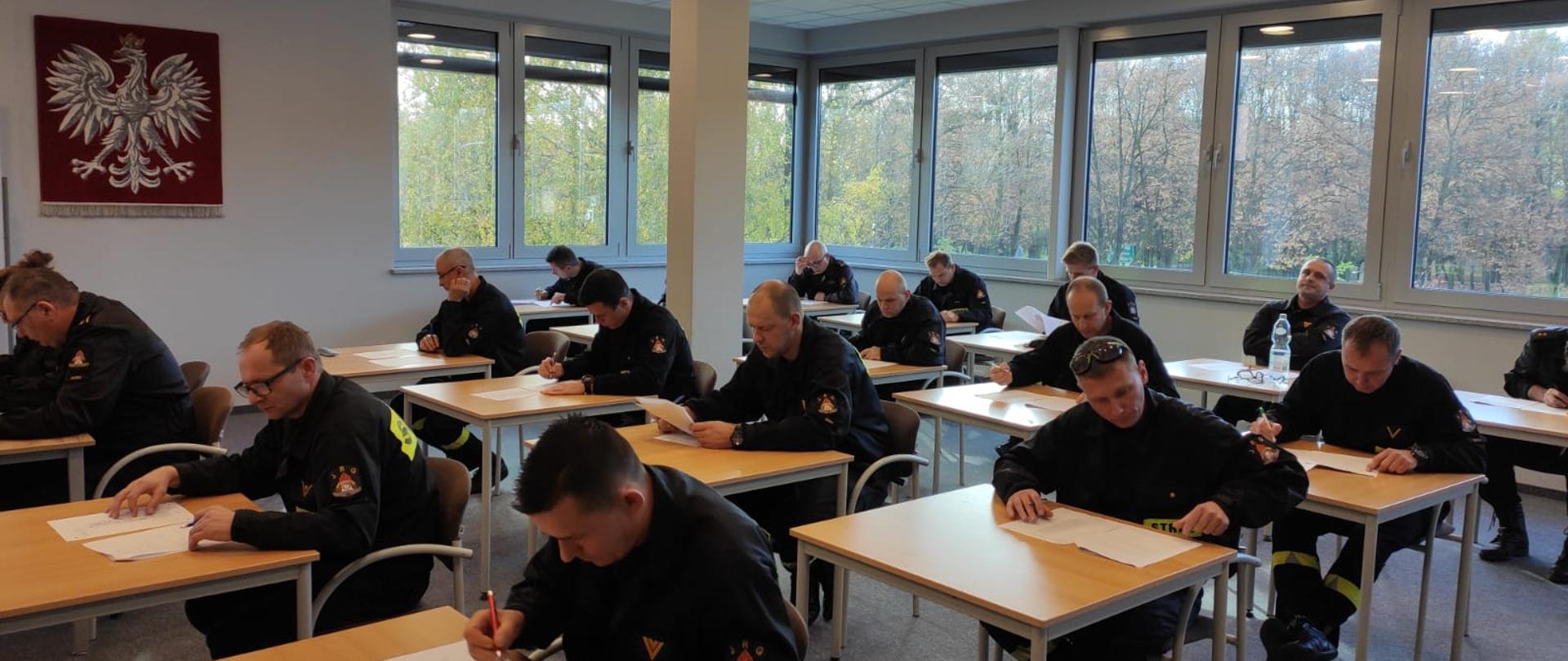 Na zdjęciu funkcjonariusze przy stolikach na świetlicy podczas rozwiązywania testu z wiedzy teoretycznej KPP na ścianie Godło Polski