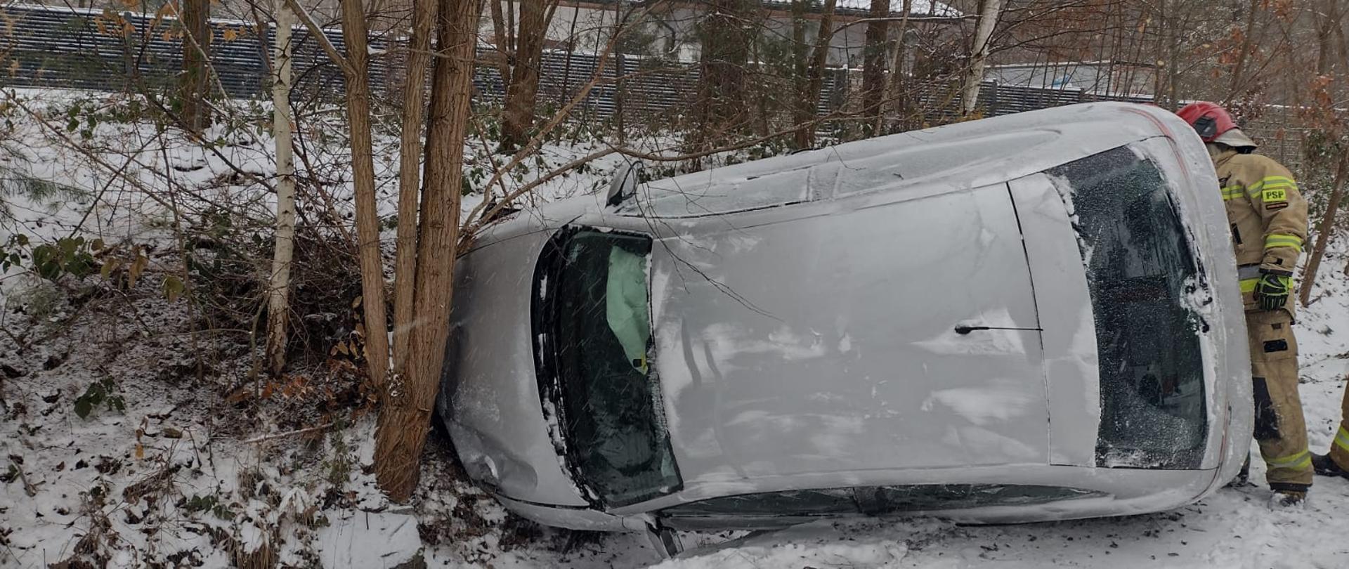 W porze dziennej leży na boku auto osobowe oparte o drzewo. Z autem widać strażaka. W tle aura zimowa - śnieżnie