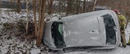 W porze dziennej leży na boku auto osobowe oparte o drzewo. Z autem widać strażaka. W tle aura zimowa - śnieżnie