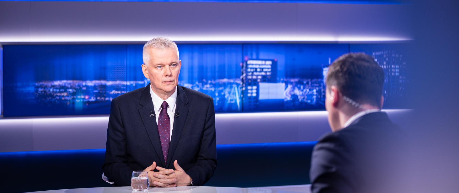 Zdjęcie przedstawia studio telewizyjne. Przy białym stole siedzą dwie osoby - po lewej minister Tomasz Siemoniak, a po prawej redaktor Piotr Witwicki (siedzący tyłem). Na stole przed ministrem stoi szklanka z wodą. W tle widać ekrany z panoramą nocnego miasta z oświetlonymi budynkami.