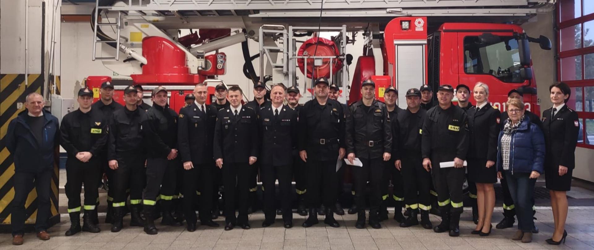 Na zdjęciu znajdują się strażacy ustawieni na garażu na tle samochodu pozrniczego