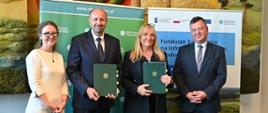 Na zdjęciu na tle banera z logo Narodowego Funduszu Ochrony Środowiska i Gospodarki Wodnej stoi od lewej: koordynator umowy Anna Siwek, Zastępca Prezesa Narodowego Funduszu Ochrony Środowiska i Gospodarki Wodnej Robert Gajda i prezes Przedsiębiorstwo Wodociągów i Kanalizacji "WODNIK" Katarzyna Wierska-Kuberska oraz Zastępca Dyrektora Departamentu Adaptacji do Zmian Klimatu Robert Markiewicz. Prezes Gajda i Prezes Wierska-Kuberska trzymają w dłoniach podpisaną umowę. 