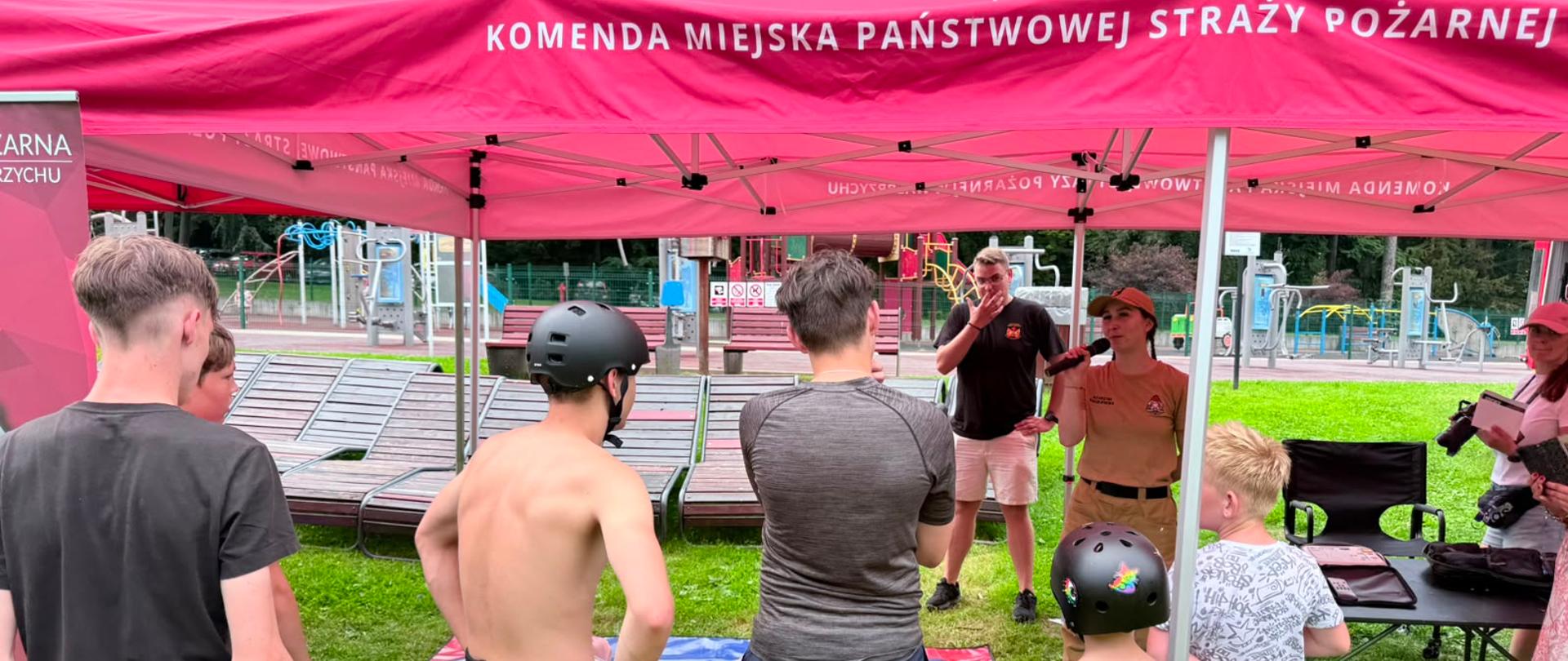 Widok spod różowego namiotu na trawiastą powierzchnię, gdzie strażaczka z mikrofonem prowadzi warsztaty pierwszej pomocy dla grupy dzieci. Chłopcy i dziewczynka w letnich strojach sportowych, niektórzy w kaskach na rolki, stoją w półkolu i słuchają, patrząc w stronę leżących na niebieskiej macie fantomów do resuscytacji. W tle widać plac zabaw, ławki parkowe oraz inne osoby przyglądające się wydarzeniu.