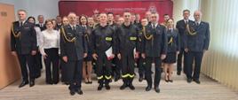 Nowo przyjęci strażacy stoją z komendantami oraz pracownikami komendy na świetlicy po ślubowaniu. Z tyłu widać baner komendy. 