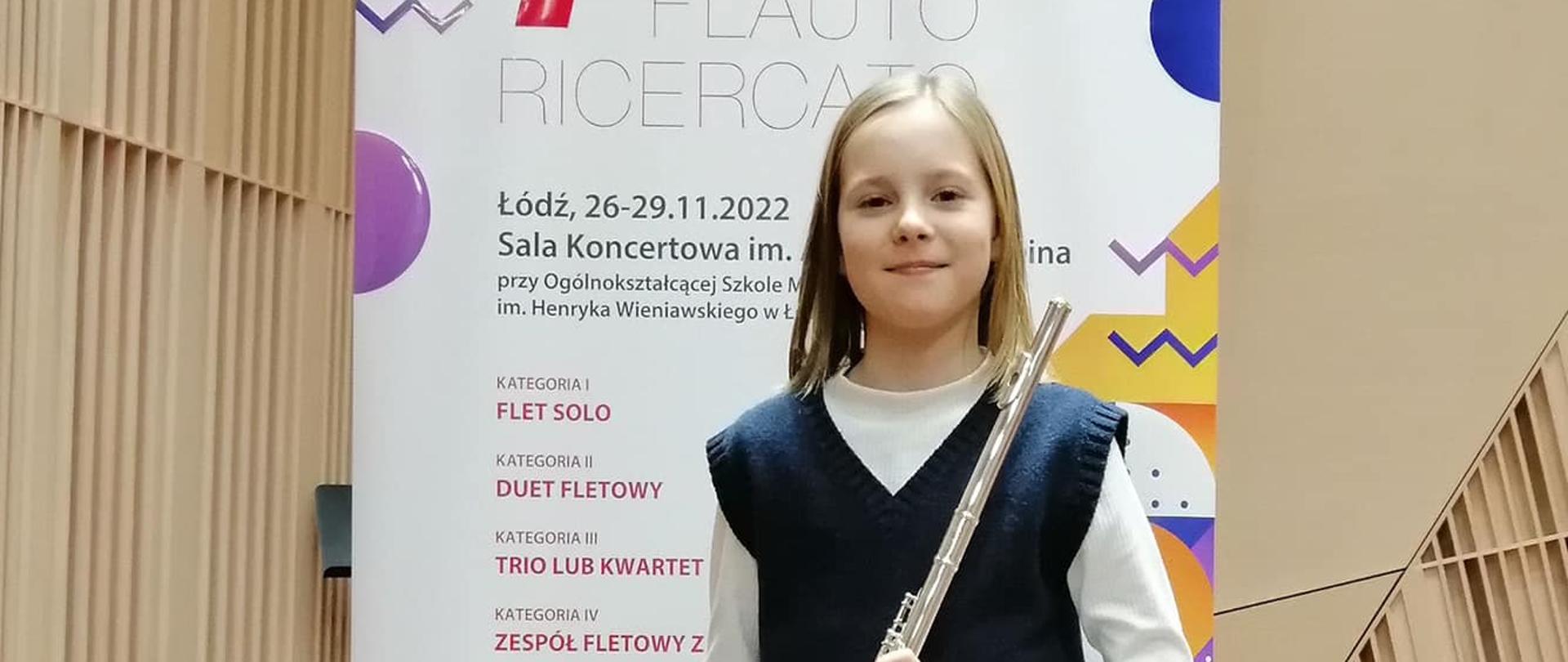 Zdjęcie przedstawia uczennicę stojącą na tle baneru konkursu, w ręku trzyma flet poprzeczny.