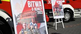 Zdjęcie wykonane podczas konferencji prasowej promującej wydarzenie "bitwa o remizy" sfinansowania przez rząd termomodernizacji i doposażenia remiz dla gmin o najwyższej frekwencji wyborczej. Na zdjęciu ustawione są pojazdy ratowniczo - gaśniczy. Pojazdy w kolorze czerwonym ze srebrnymi oraz białymi elementami zabudowy. Przed pojazdami na sztalugach ustawione są również plakat promujący to wydarzenie. Na plakacie widać trzech strażaków ubranych w ubrania specjalne w kolorze żółtym. Dwóch z nich na głowach ma założone białe hełmy. Za nimi w tle widać remizę strażacką w kolorze czerwonym. Nad nią znajduje się napis w kolorze białym bitwa o remizy 2023.