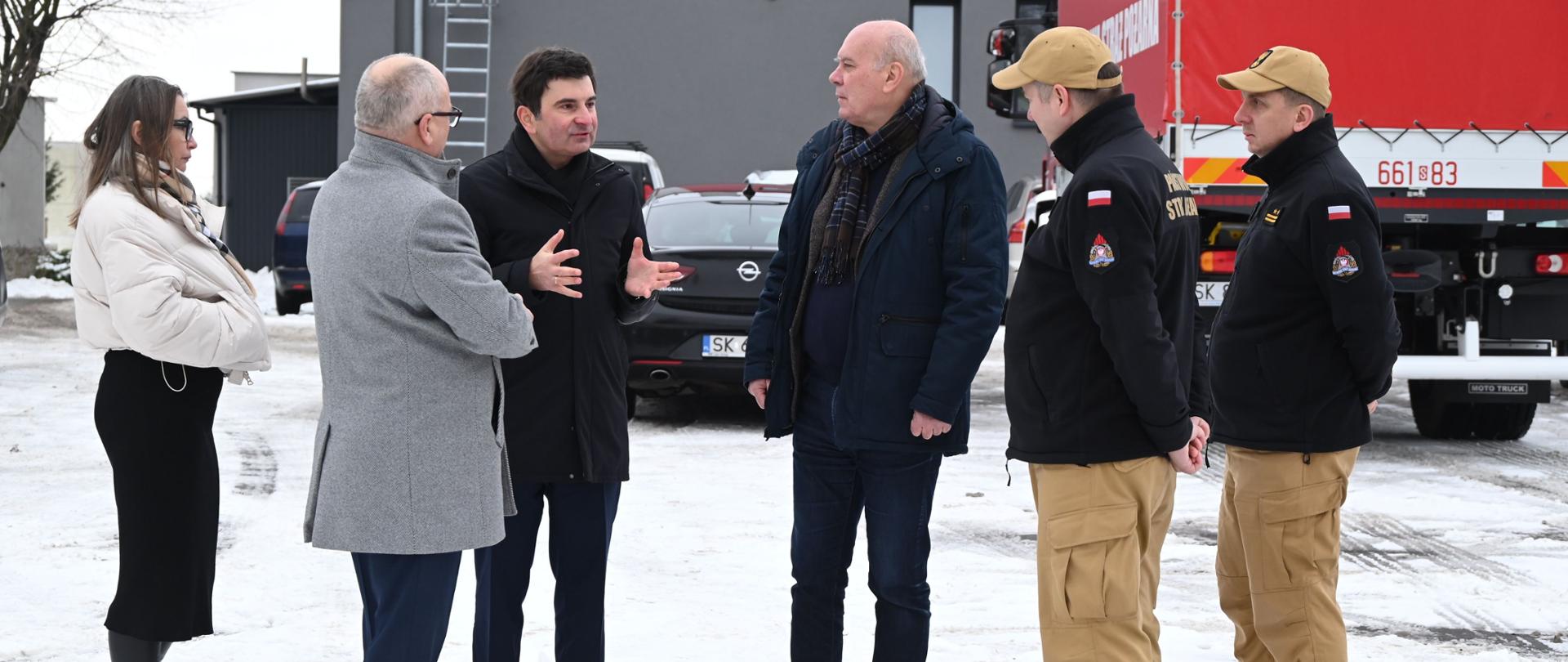 Na zaśnieżonym placu przed budynkiem przemysłowym stoi grupa siedmiu osób podczas rozmowy służbowej. Wśród nich znajdują się komendant powiatowy, wojewoda śląski, poseł, starosta oraz wicestarosta, ubrani w zimowe płaszcze. Dwóch funkcjonariuszy w umundurowaniu PSP stoi z boku i przysłuchuje się rozmowie. W tle widoczne są zaparkowane samochody oraz czerwony samochód ciężarowy.