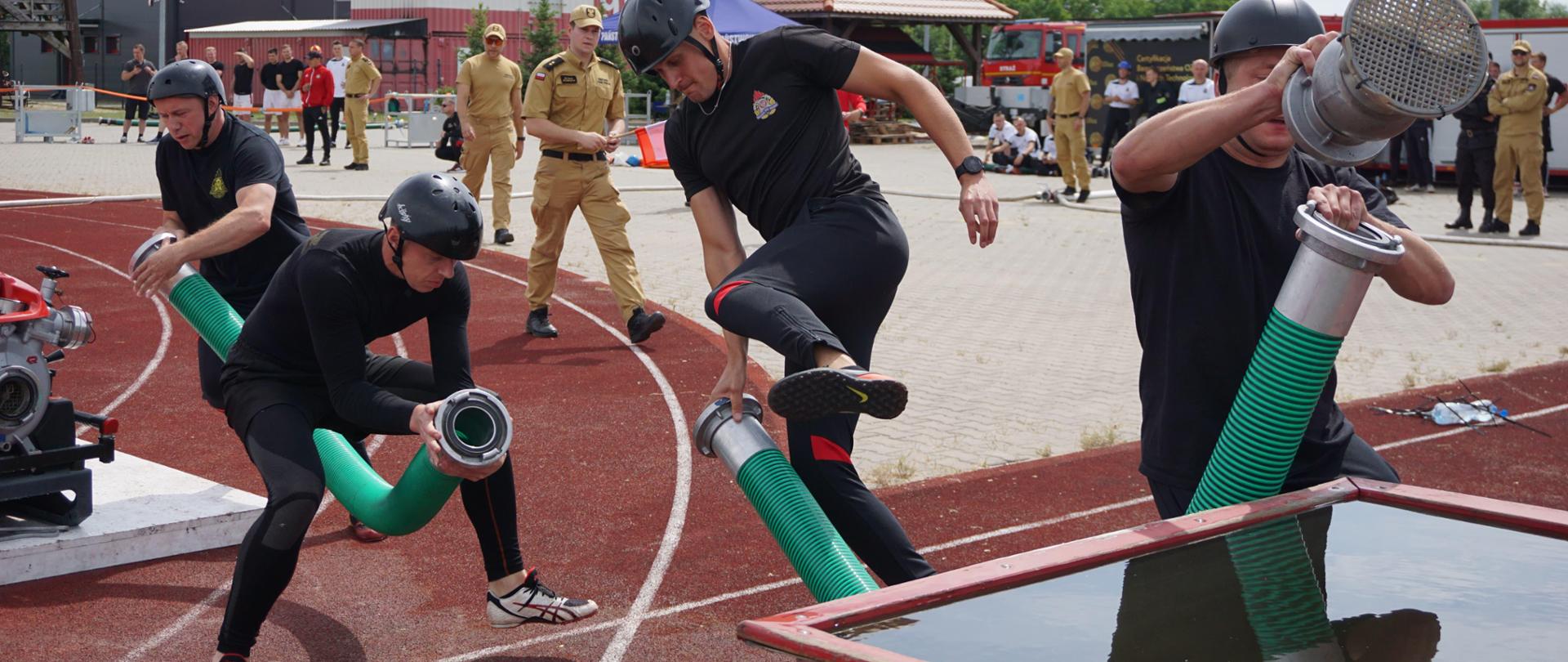 Czterech strażackich zawodników sportowych sprawiają linię ssawną. W tle obserwatorzy, dalej obiekty treningowe ośrodka szkolenia.