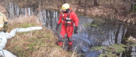 Na zdjęciu strażak w czerwonym kombinezonie rozstawia zaporę przeciwolejową na cieku wodnym.
