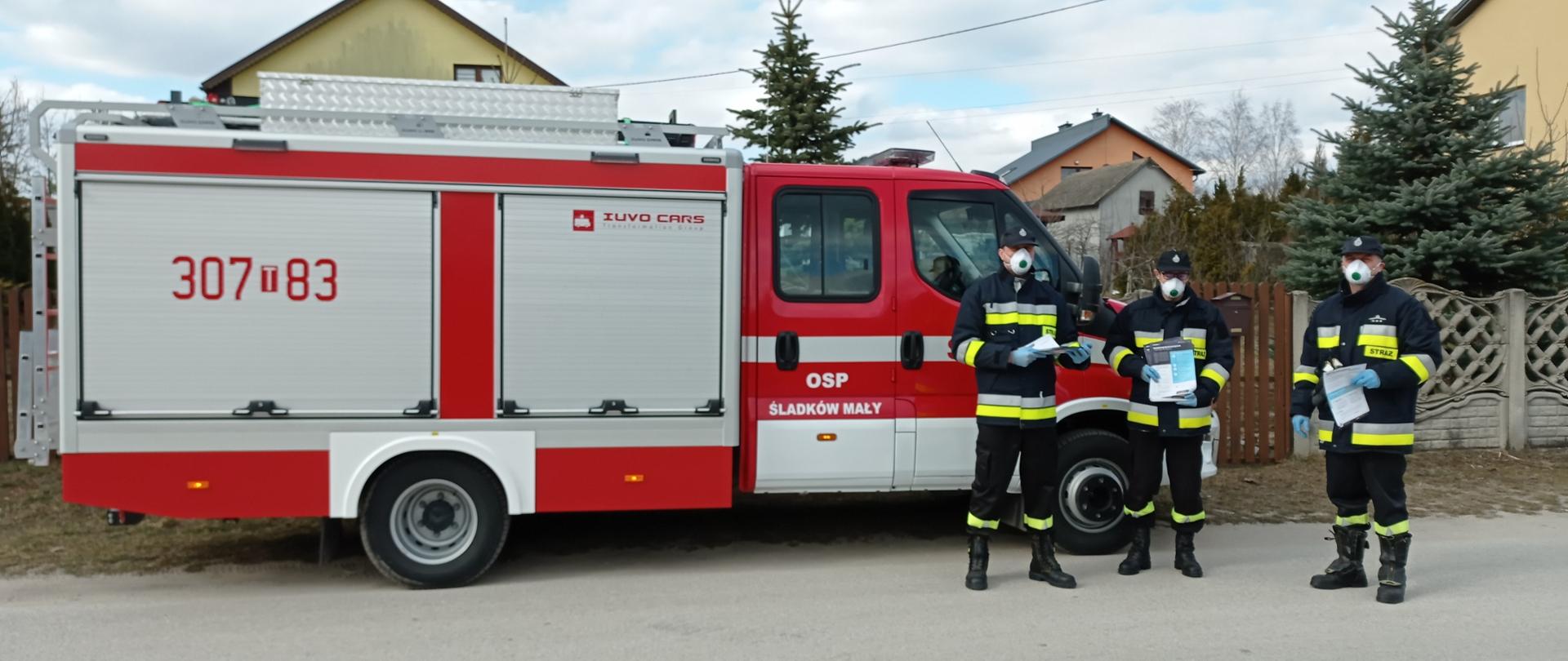 Zdjęcie przedstawia samochód OSP Śladków Mały oraz strażaków z tej jednostki stojących z prawej strony samochodu i trzymających ulotki dot. szczepień