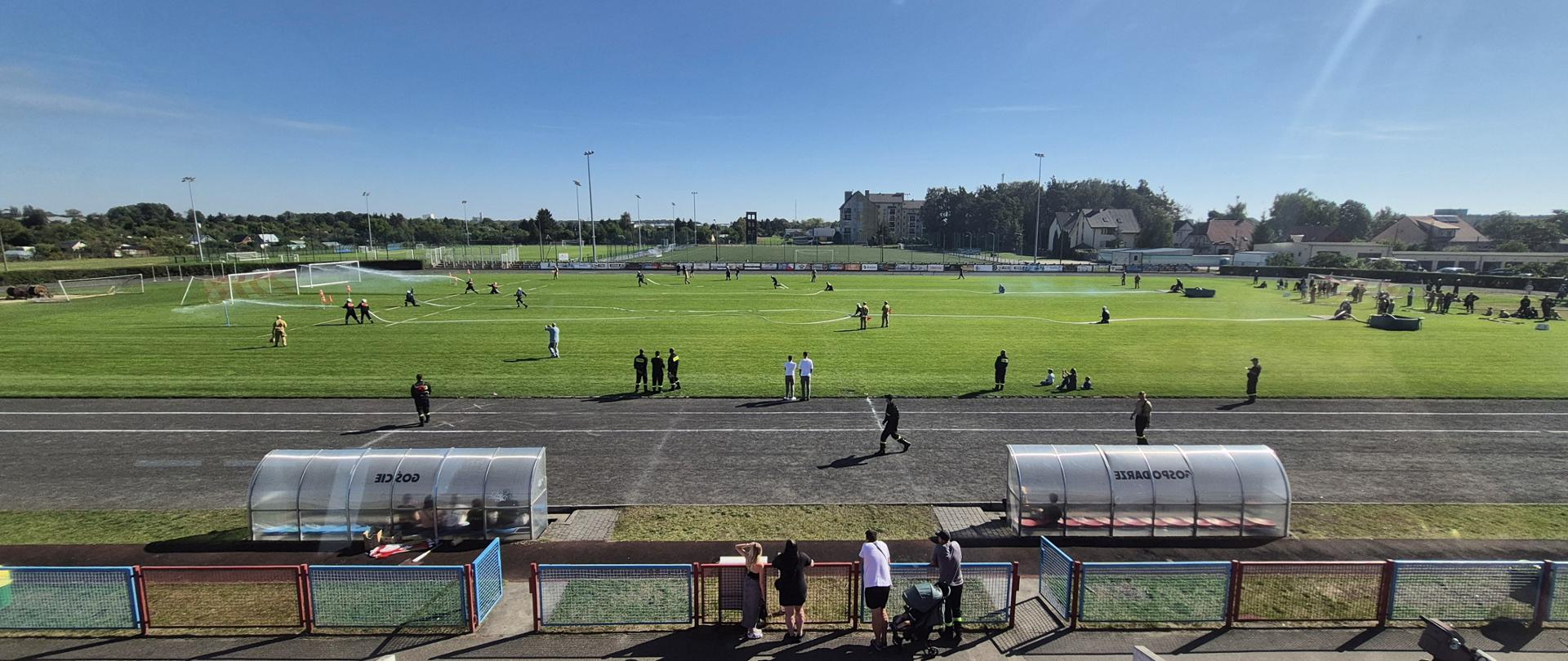 Na zdjęciu widać stadion z zieloną murawą, na której odbywają się zawody strażackie. Kilka drużyn strażaków w umundurowaniu bierze udział w konkurencjach z użyciem węży strażackich, przez które tryska woda. Na bieżni wokół boiska stoją sędziowie, organizatorzy i obserwatorzy. W tle widoczne są trybuny, zabudowania mieszkalne oraz drzewa. Na pierwszym planie, przy ogrodzeniu, stoi grupa widzów oglądających zmagania, a obok ustawione są wózki dziecięce. Atmosfera wydarzenia jest sportowa i rekreacyjna, a pogoda słoneczna.