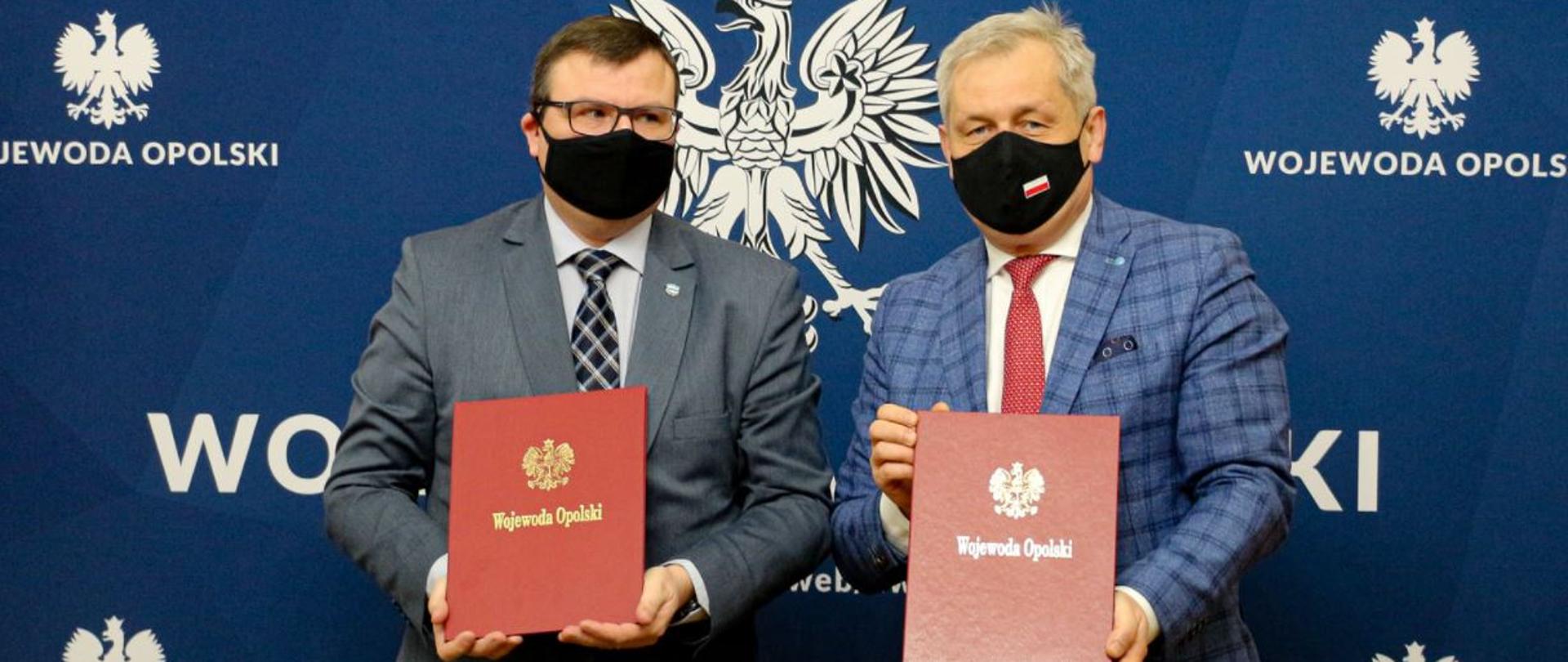 Na zdjęciu wojewoda opolski, stojący obok dyrektora PGW WP RZGW Gliwice. W dłoniach trzymają czerwone teczki. W tle granatowa ścianka z napisem Wojewoda Opolski. 