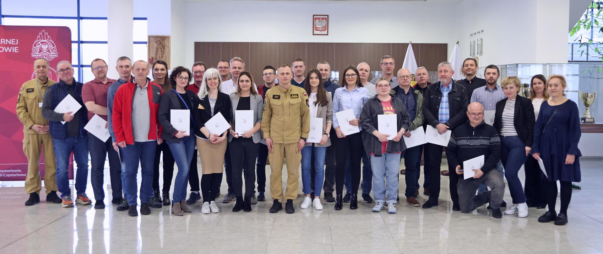 Pamiątkowe zdjęcie grupowe uczestników szkolenia aktualizującego inspektorów ochrony przeciwpożarowej (27 osób) w towarzystwie Zastępcy Komendanta Centralnej Szkoły PSP, kobiety oraz funkcjonariusza CS PSP
