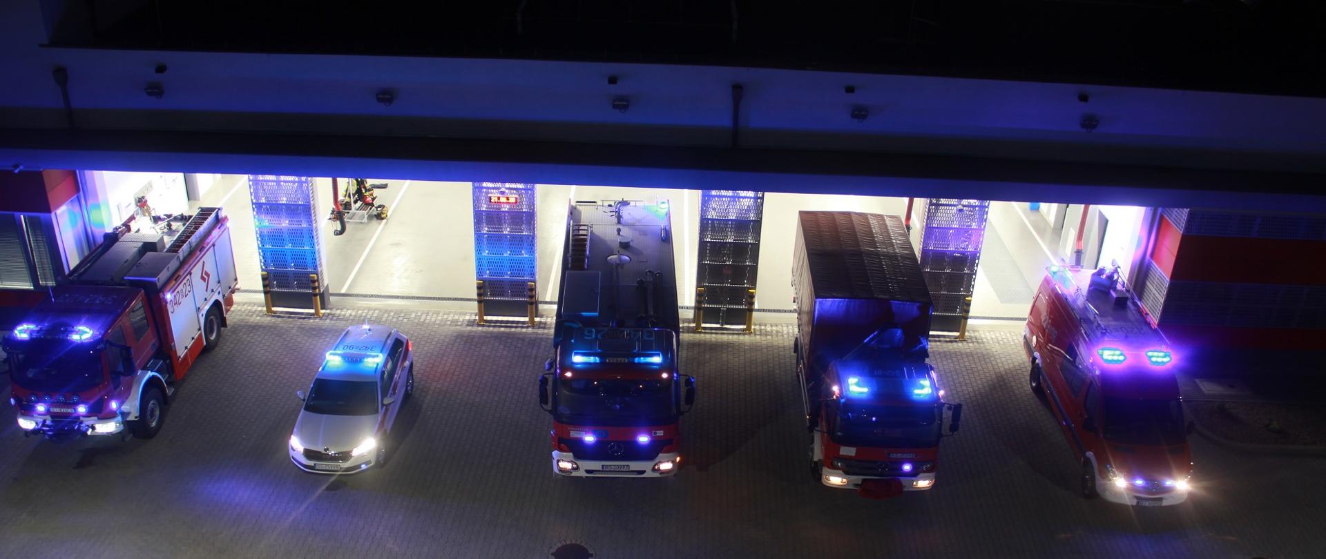 Na zdjęciu widać w nocy przed bramami garaży 5 samochodów strażackich z włączonymi sygnałami świetlnymi. 