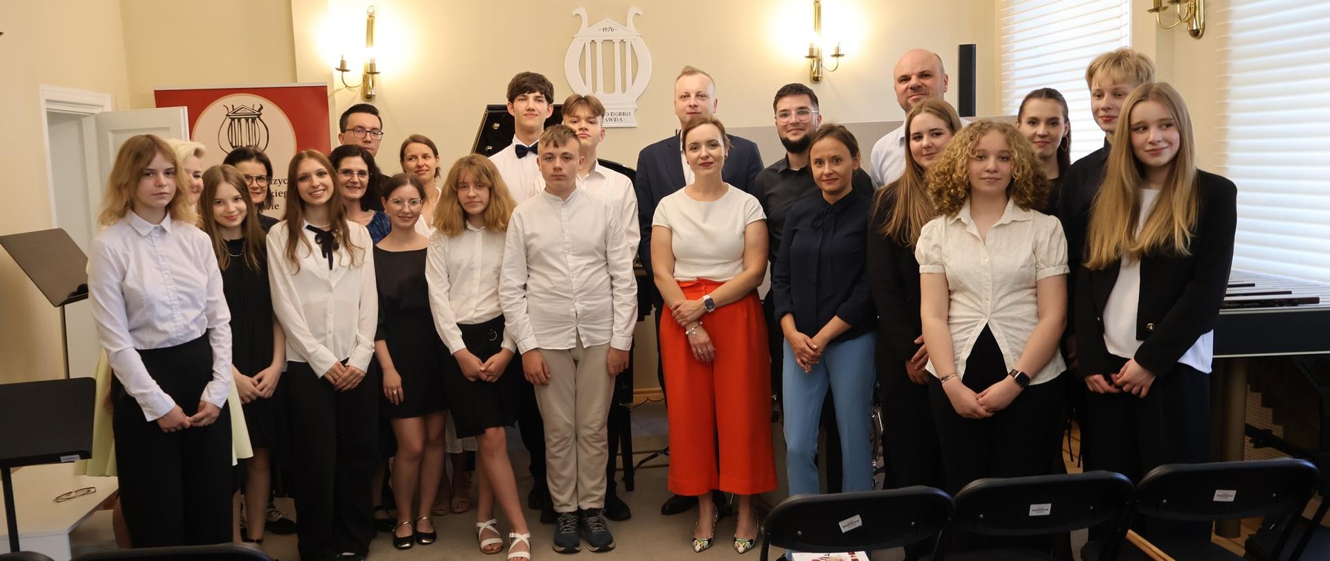 Uczniowie i nauczyciele stojący w auli szkoły.