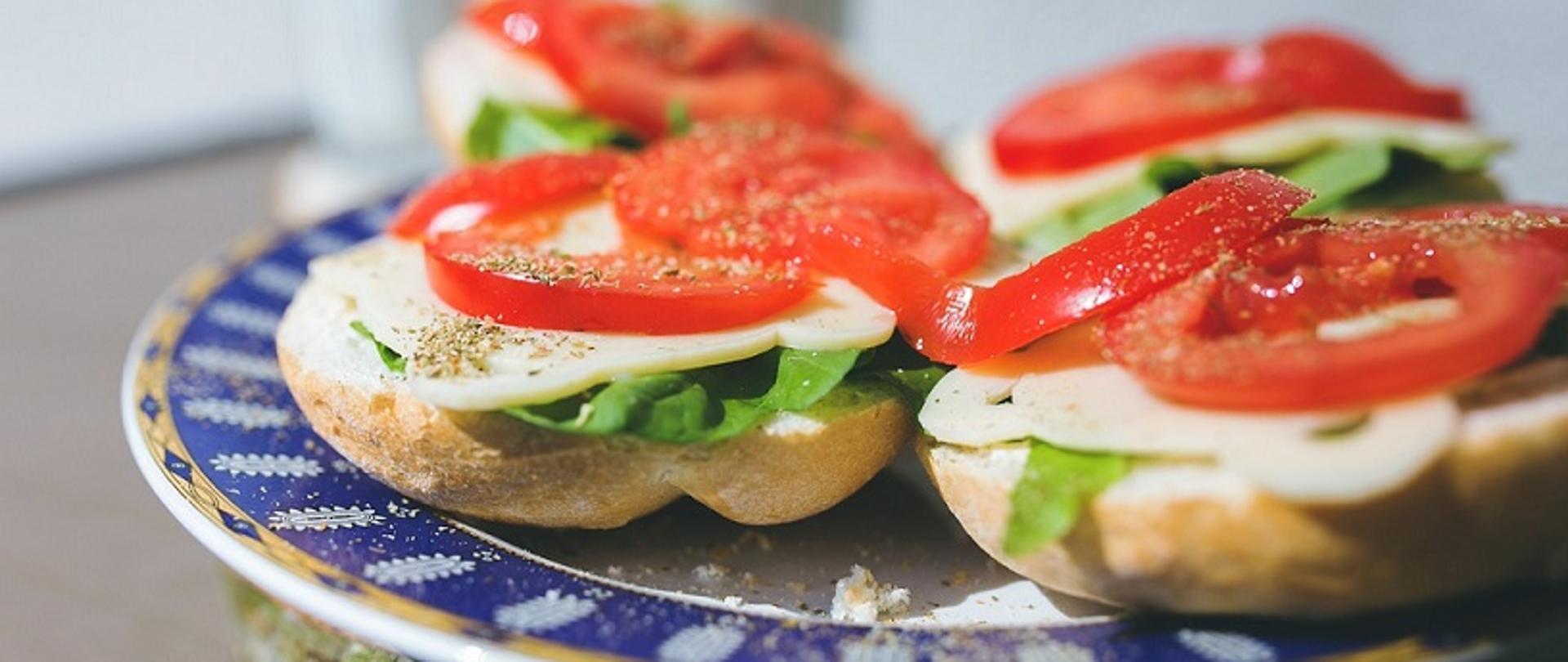 na talerzyku śniadaniowym kanapki z serem żółtym, sałatą, pomidorami, posypane ziołami
