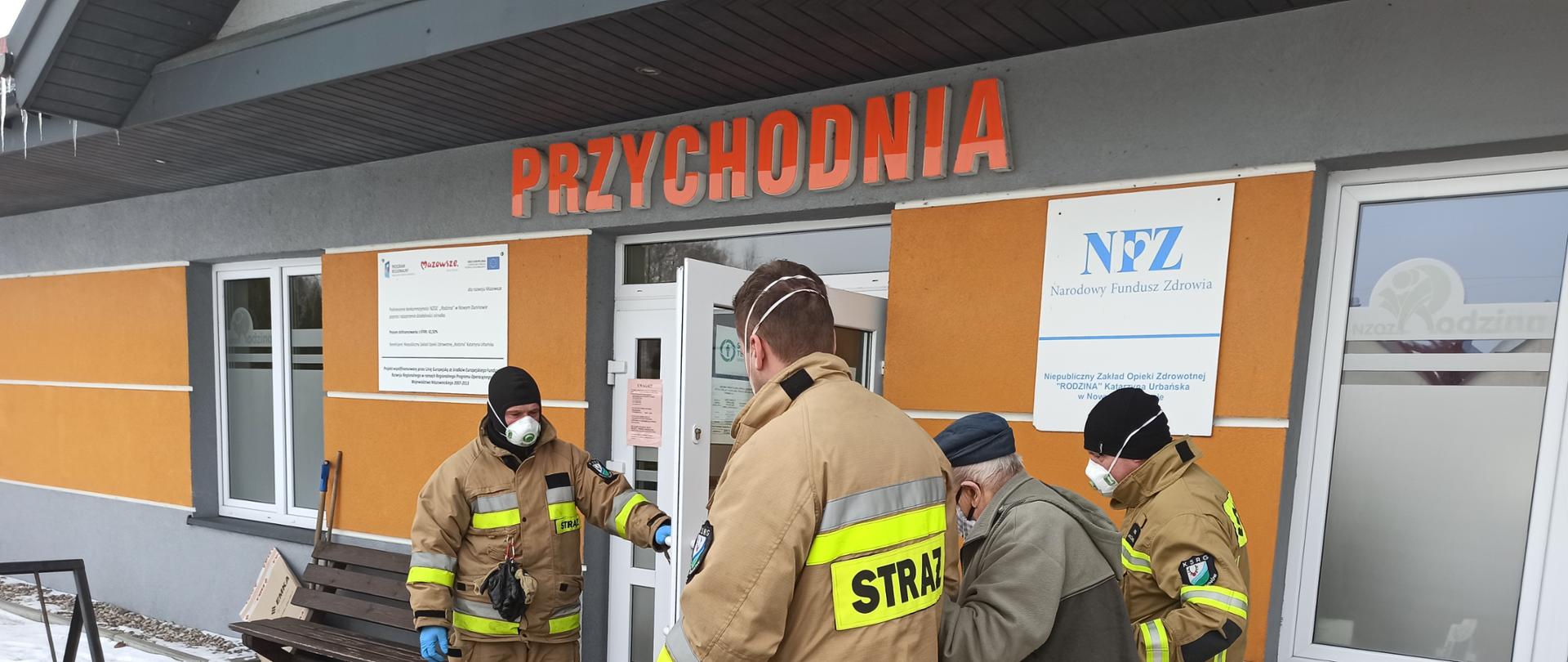 Na zdjęciu strażacy OSP Nowy Duninów pomagają seniorowi wejść do ośrodka zdrowia w Nowym Duninowie. Dwóch strażaków prowadzi seniora pod ręce, a trzeci otwiera drzwi do ośrodka.