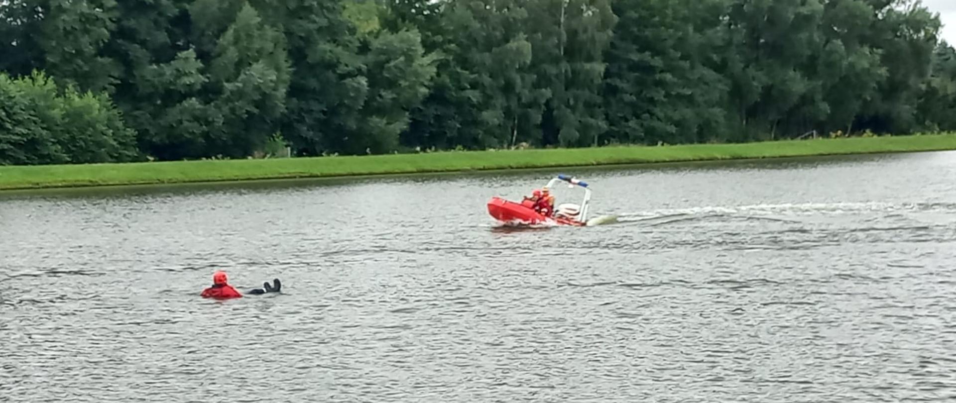 Zdjęcie przedstawia dwóch strażaków na czerwonej łodzi płynących do osoby poszkodowanej znajdującej się w wodzie, która zabezpieczona jest w specjalne ubranie, kamizelkę utrzymującą na wodzie oraz kask. W tle znajdują się drzewa za zbiornikiem wodnym.