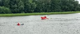 Zdjęcie przedstawia strażaków na czerwonej łodzi płynących do osoby poszkodowanej znajdującej się w wodzie, która zabezpieczona jest w specjalne ubranie, kamizelkę utrzymującą na wodzie oraz kask. W tle znajdują się drzewa za zbiornikiem wodnym.