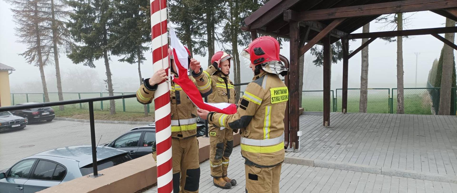 Pora dzienna, Na placu przy maszcie trzech strażaków w umundurowaniu specjalnym zakłada flagę państwową na maszt. 