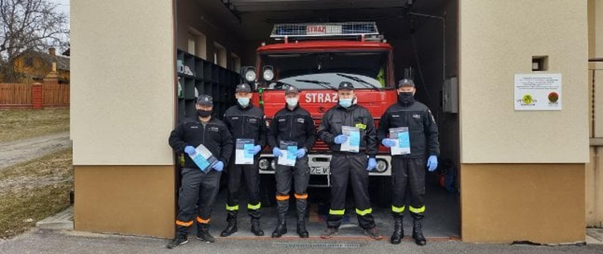 Na zdjęciu widocznych jest 5 mężczyzn - druhów OSP z ulotkami w rękach. Zabezpieczeni są w maseczki na twarzy oraz rękawiczki. Znajdują się świetle bramy garażowej jednostki OSP,. Wewnątrz garażu znajduje się czerwony samochód gaśniczy.