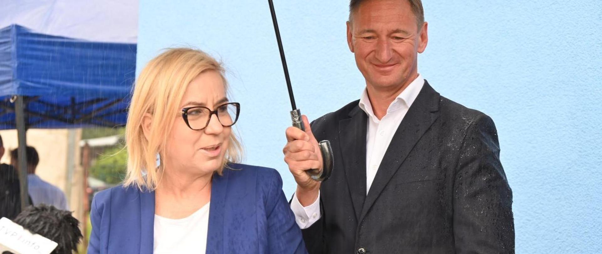 Minister Paulina Hennig-Kloska during the opening of the first building to undergo comprehensive modernisation under the Multi-Family Buildings in Rural Areas Modernisation Programme in Rymań. In the photograph, the Minister is standing and smiling while speaking into a microphone. Behind her, a man is holding an open umbrella over her.