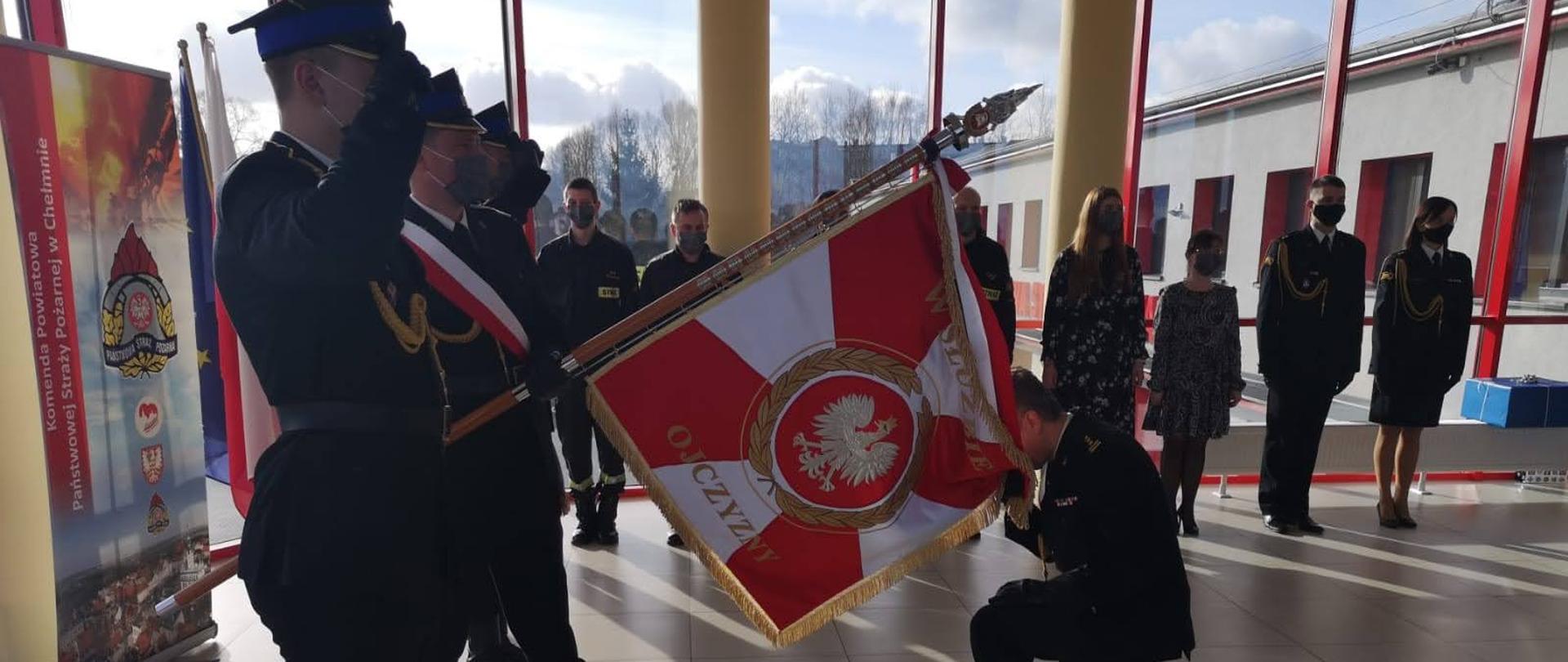 Na zdjęciu w sali sztabowej Komendy Powiatowej PSP w Chełmnie znajdują się funkcjonariusze i pracownicy cywilni komendy podczas uroczystej zbiórki z okazji odejścia za zaopatrzenie emerytalne st. bryg. Janusza Pietraka, który przyklęka przy sztandarze komendy żegnając się z nim. 