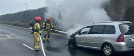 Pożar samochodu osobowego. Pożarem objęta komora silnika. Na autem unosi się gęstym dym. Strażacy podają pianę gaśniczą na palący się pojazd