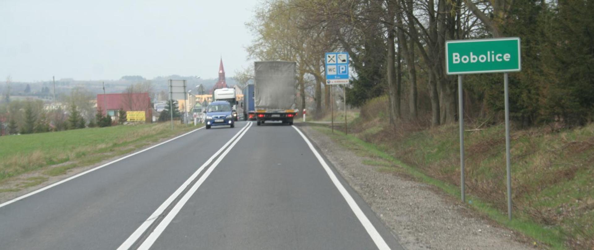 Dwupasmowa droga w terenie podmiejskim, z podwójną linią ciągłą na jezdni. Na drodze jadą samochody, w tym ciężarówka. Po prawej stronie stoi zielony znak miejscowości z napisem „Bobolice”, a dalej znak informujący m.in. o parkingu i toalecie. W tle widoczne domy, drzewa oraz wieża kościoła.