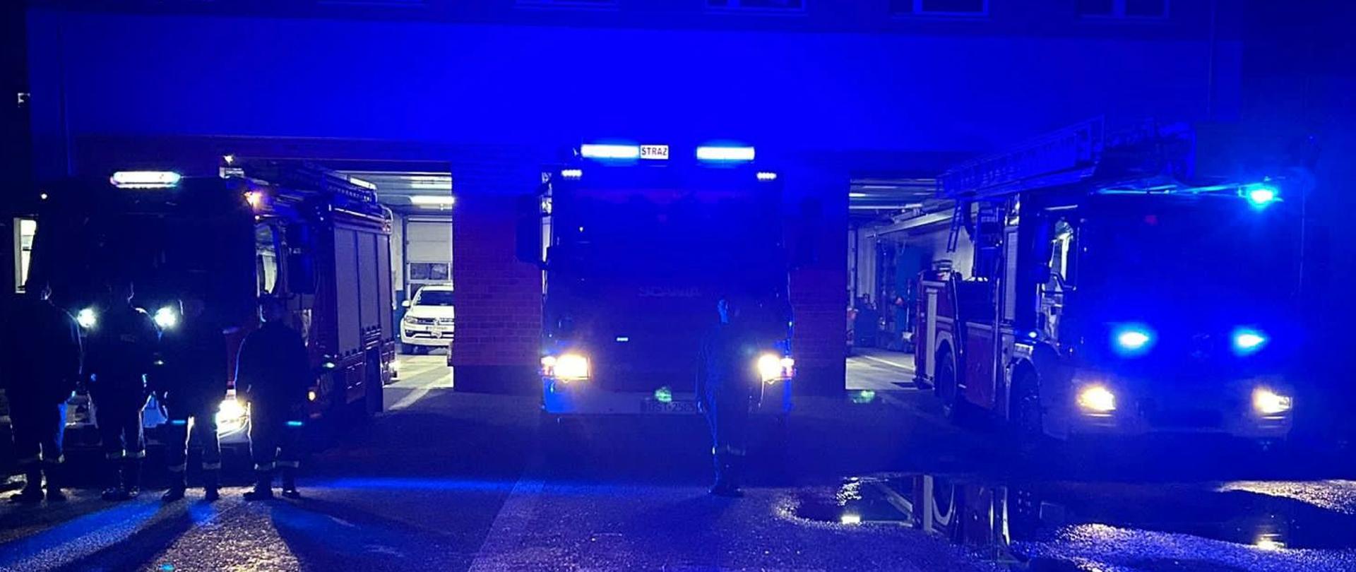 Na zdjęciu znajdują się wozy strażackie z włączonymi światłami oraz światłami wywoławczymi, wokoło panuje mrok