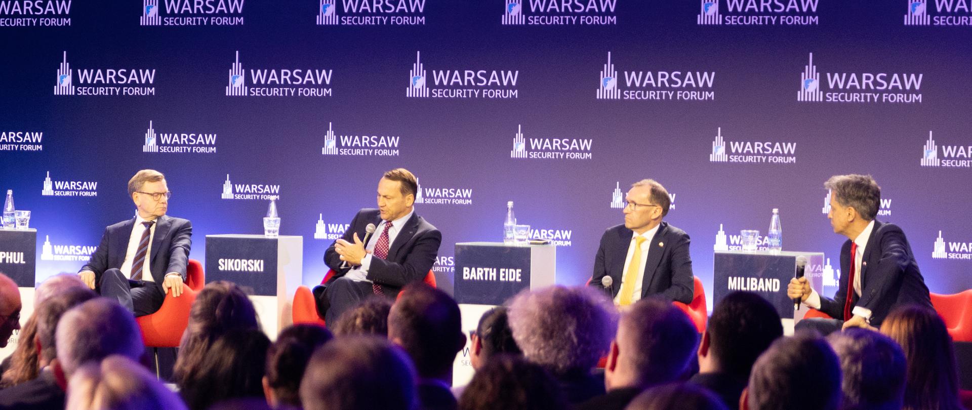 Radosław Sikorski, wicepremier i minister spraw zagranicznych Polski, bierze udział w dyskusji panelowej podczas Warsaw Security Forum. Siedzi na scenie wśród pozostałych panelistów, a jego wizerunek jest wyświetlany na dużym ekranie po lewej stronie. W tle widoczne są logotypy wydarzenia oraz fioletowe i niebieskie oświetlenie sceniczne.