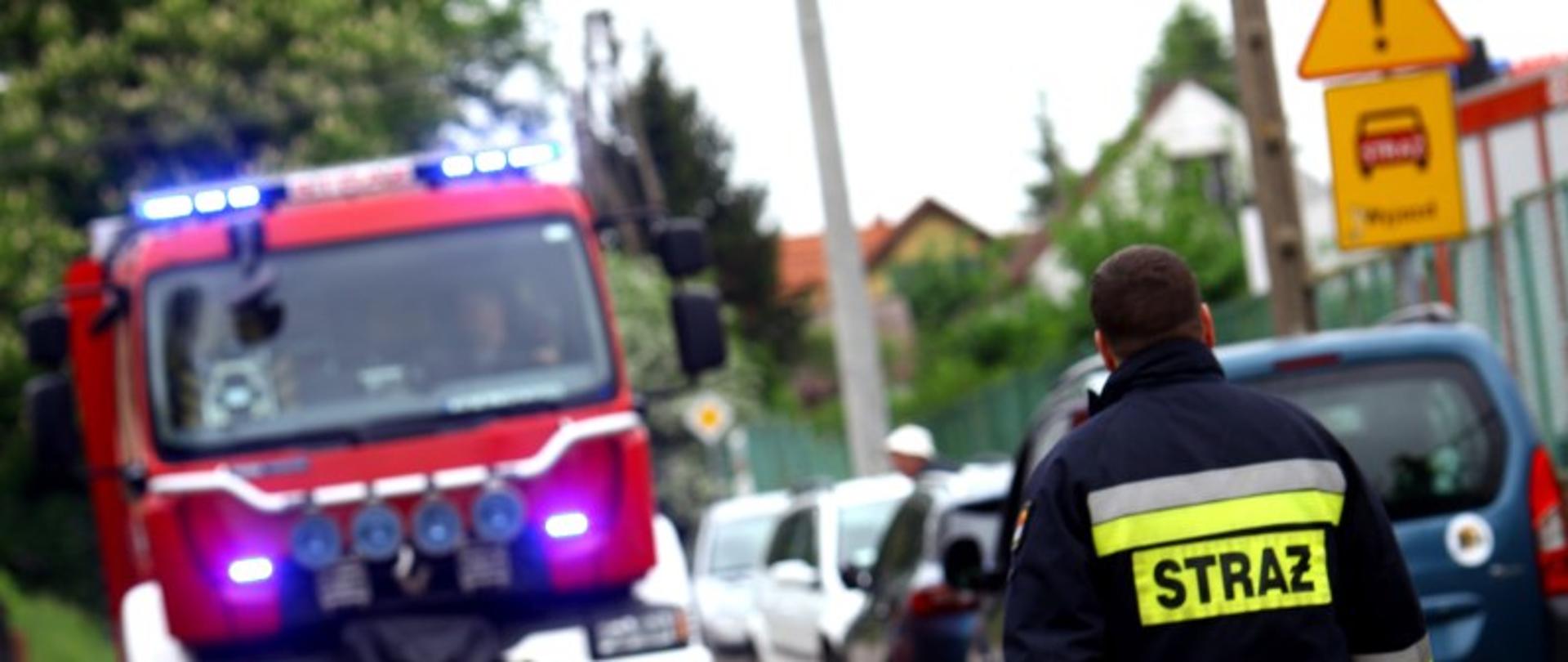 Zdjęcie przedstawia strażaka idącego drogą, w t;e widać wóz straży pożarnej.