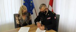 Na zdjęciu przedstawicielka Firmy CEDROB oraz Kujawsko-Pomorski Komendant Wojewódzki PSP st. bryg. Jacek Kaczmarek, siedzą przy stole i podpisują dokumenty. W tle flaga Polski i Unii Europejskiej.