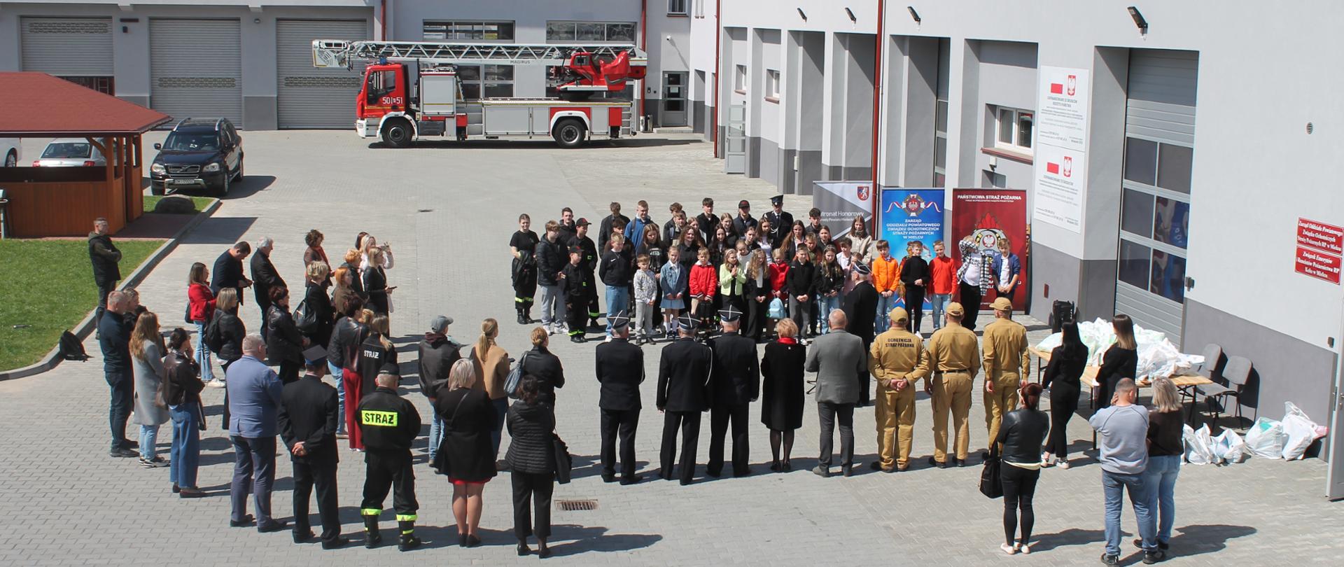Szerokie ujęcie z góry na plac przed komendą. Uczestnicy i goście formują półokrąg podczas oficjalnej ceremonii zakończenia konkursu. Na środku stoją strażacy i organizatorzy. Po prawej stronie widoczny stolik z nagrodami zapakowanymi w białe torby.