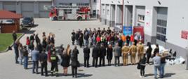 Szerokie ujęcie z góry na plac przed komendą. Uczestnicy i goście formują półokrąg podczas oficjalnej ceremonii zakończenia konkursu. Na środku stoją strażacy i organizatorzy. Po prawej stronie widoczny stolik z nagrodami zapakowanymi w białe torby.