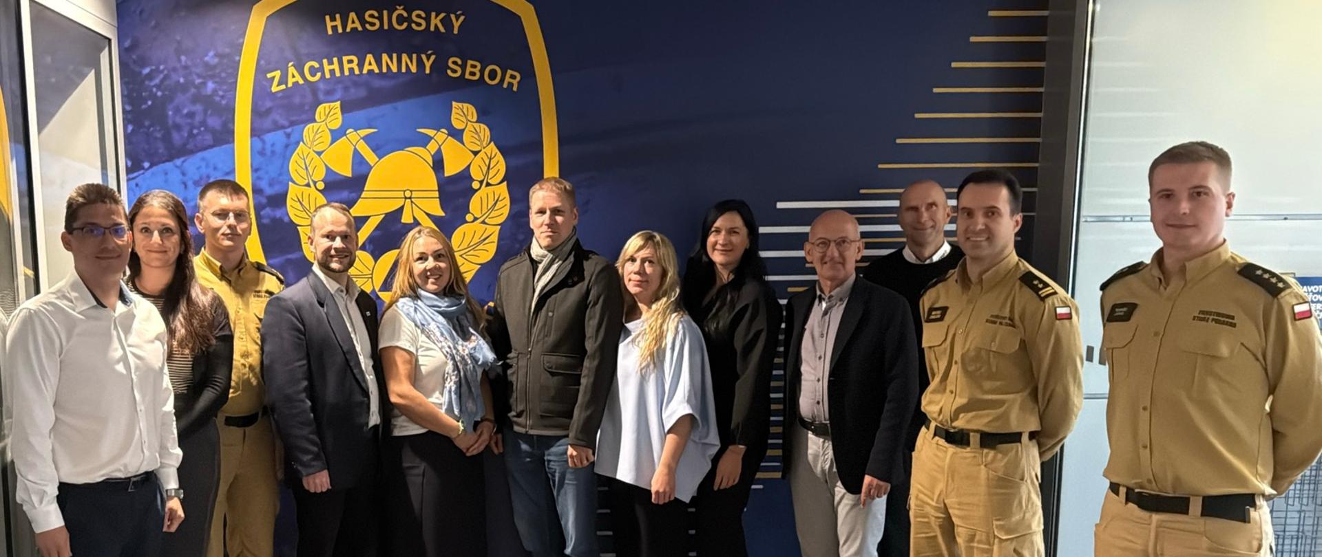 Grupa kilkunastu osób pozuje do wspólnego zdjęcia wewnątrz budynku czeskiej straży pożarnej. Wśród uczestników są funkcjonariusze PSP w mundurach oraz osoby w ubraniach cywilnych. 
