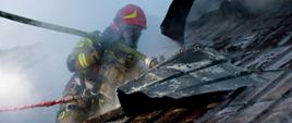 Zdjęcie wykonane podczas działań ratowniczo – gaśniczych w budynku mieszkalnym. Na zdjęciu widać dach budynku kryty blachą. Na dachu budynku znajduje się jeden strażak. Ubrany jest w ubrania specjalne w kolorze piaskowym. Na głowie ma hełm w kolorze czerwonym. Na rękach rękawice ochronne. Na twarzy maskę ochronną połączoną z butą ze sprężonym powietrzem znajdującą się na ich plecach. Dodatkowo trzyma w rękach węża z którego leje wodę na płomienie wewnątrz budynku przez rozerwaną na dachu blachę. 