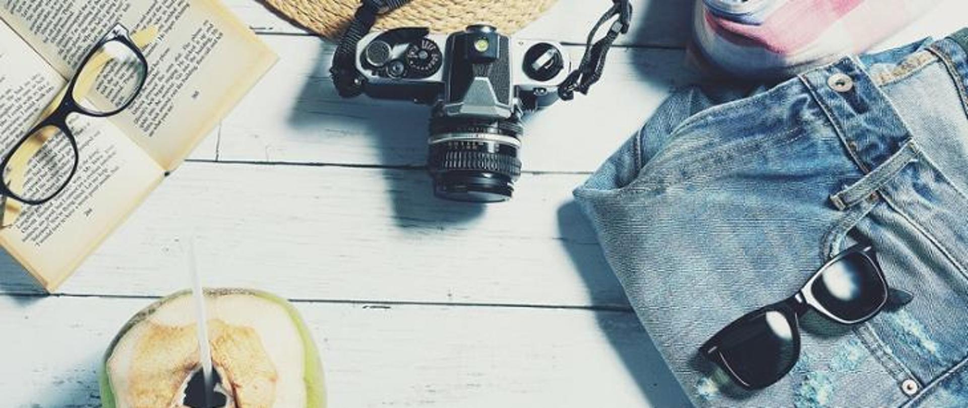 Grafika przedstawia książkę, okulary, kapelusz, letnią odzież, aparat i kokosa, położone na deskach