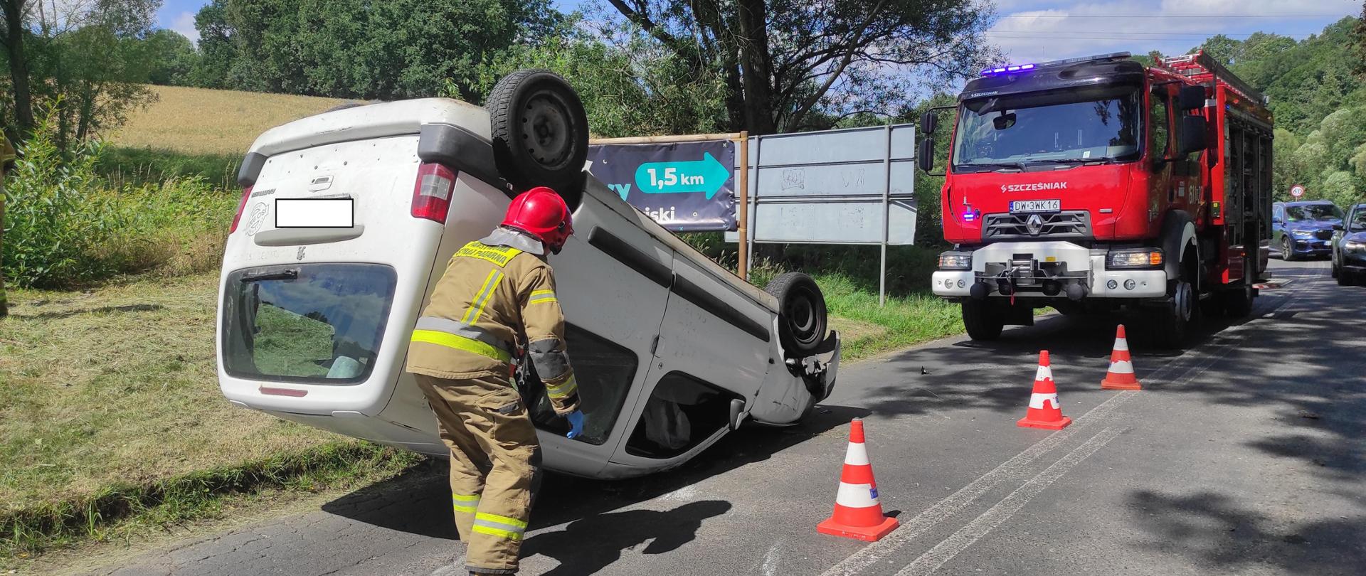 samochód osobowy leży na dachu na pasie drogowym, obok niego stoi strażak, na drodze stoi również czerwony pojazd pożarniczy