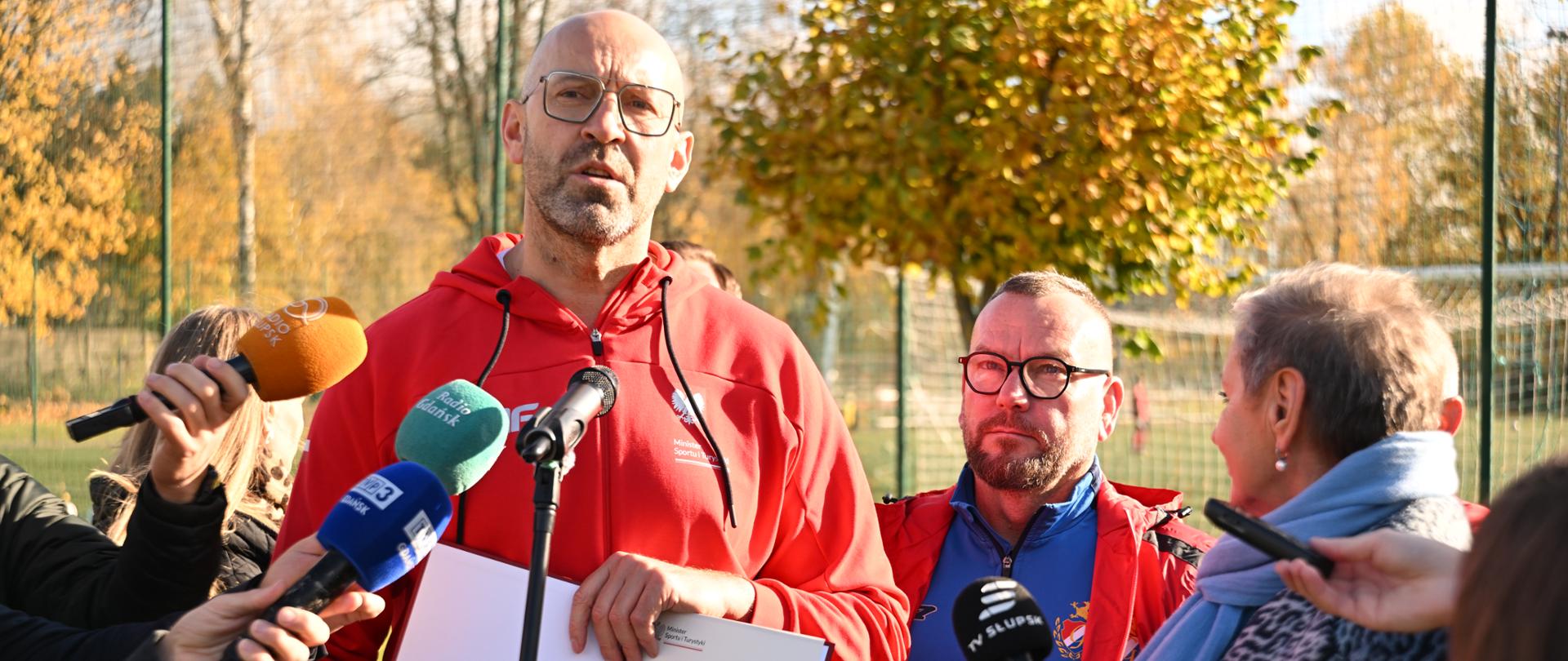 Zdjęcie przedstawiające ministra sportu i turystyki Jakuba Rutnickiego podczas konferencji prasowej. Minister trzyma w rękach decyzję o dofinansowaniu. Dookoła stoją dziennikarze z mikrofonami. Za nim z prawej strony prezes Gryfa Słupsk Mirosław Lewandowski i prezydent Słupska Krystyna Danilecka-Wojewódzka. W tle widać siatkę i drzewa.