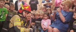 Strażak ubrany w ubranie bojowe, hełm, oraz aparat powietrzny pokazuje zgromadzonym za nim dzieciom zasadę działania kamery termowizyjnej.