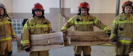 Strażacy odchodzący na emeryturę trzymają pamiątkowe tablice wykonane z drewna z przymocowanymi do nich toporami strażackimi. i napisami okolicznościowymi z podziękowaniem za służbę. 