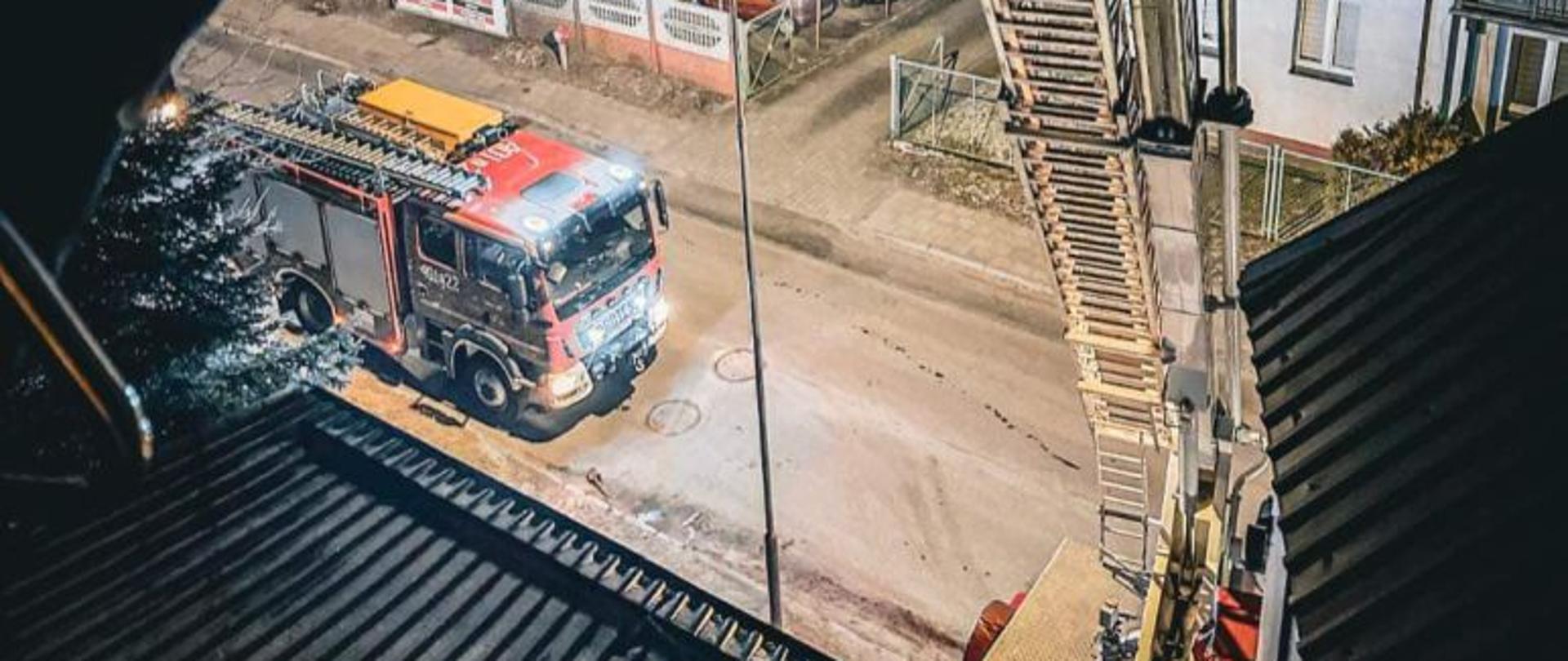 Zdjęcie zrobione z kosza podnośnika. Widać ramie podnośnika, na dole pojazdy gaśnicze. Zdjęcie wykonane w nocy.
