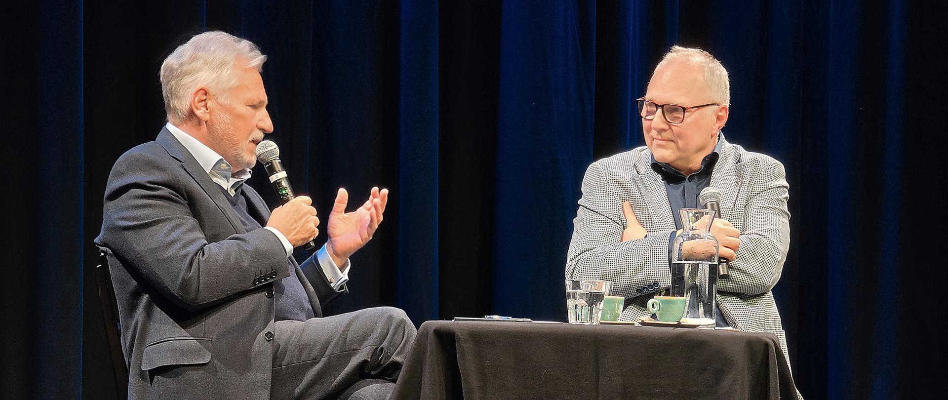 Zdjęcie przedstawia prezydenta Aleksandra Kwaśniewskiego i redaktora Jarosława Kurskiego prowadzących rozmowę na scenie. 