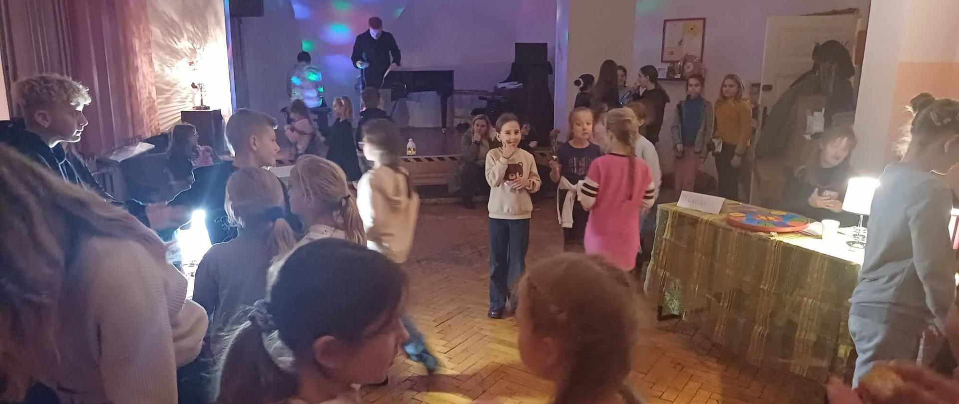 Zdjęcie zbiorowe - dzieci w sali, stół z wróżbami, na scenie w oddali DJ