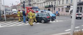 Na zdjęciu rozbity pojazd na drodze publicznej. Zdjęcie w ciągu dnia. W tle zabudowa miejska. Zachmurzenie duże. Inni uczestnicy ruchu. Funkcjonariusze PSP.