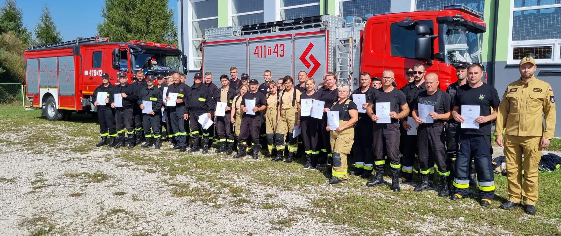 Zdjęcie przedstawia strażaków w umundurowaniu koszarowym na placu Szkoły Podstawowej w Węgleszynie. Strażacy trzymają w rękach dokumenty potwierdzające ukończenie szkolenia podstawowego. W tle dwa samochody ratowniczo-gaśnicze.