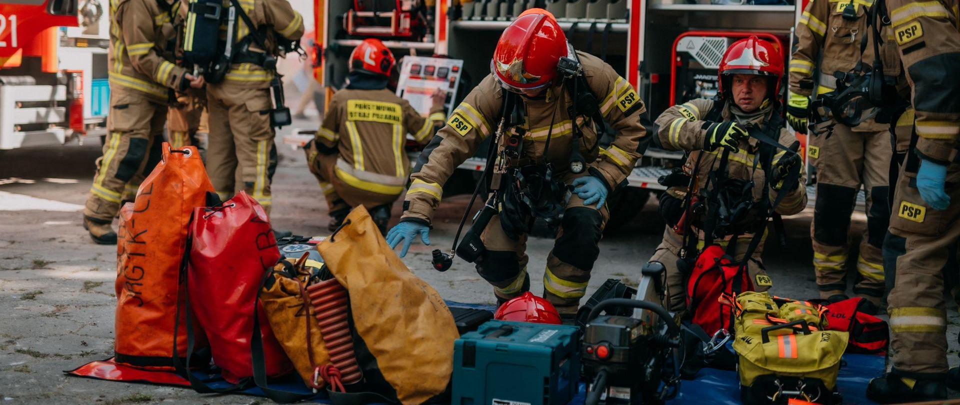 Na zdjęciu widzimy strażaków podczas przygotowania do wejścia na teren opuszczonego szpitala. Na macie ułożony sprzęt potrzebny do działań, a za nimi samochód pożarniczy z otwartymi skrytkami.