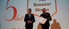 Pomorski komendant wojewódzki Państwowej Straży Pożarnej trzyma wyróżnienie obok stoi prezesem Rady Ministrów za nimi jest umieszczony baner z napisem pięć lat konwent morski Polak potrafi wykrzyknik.