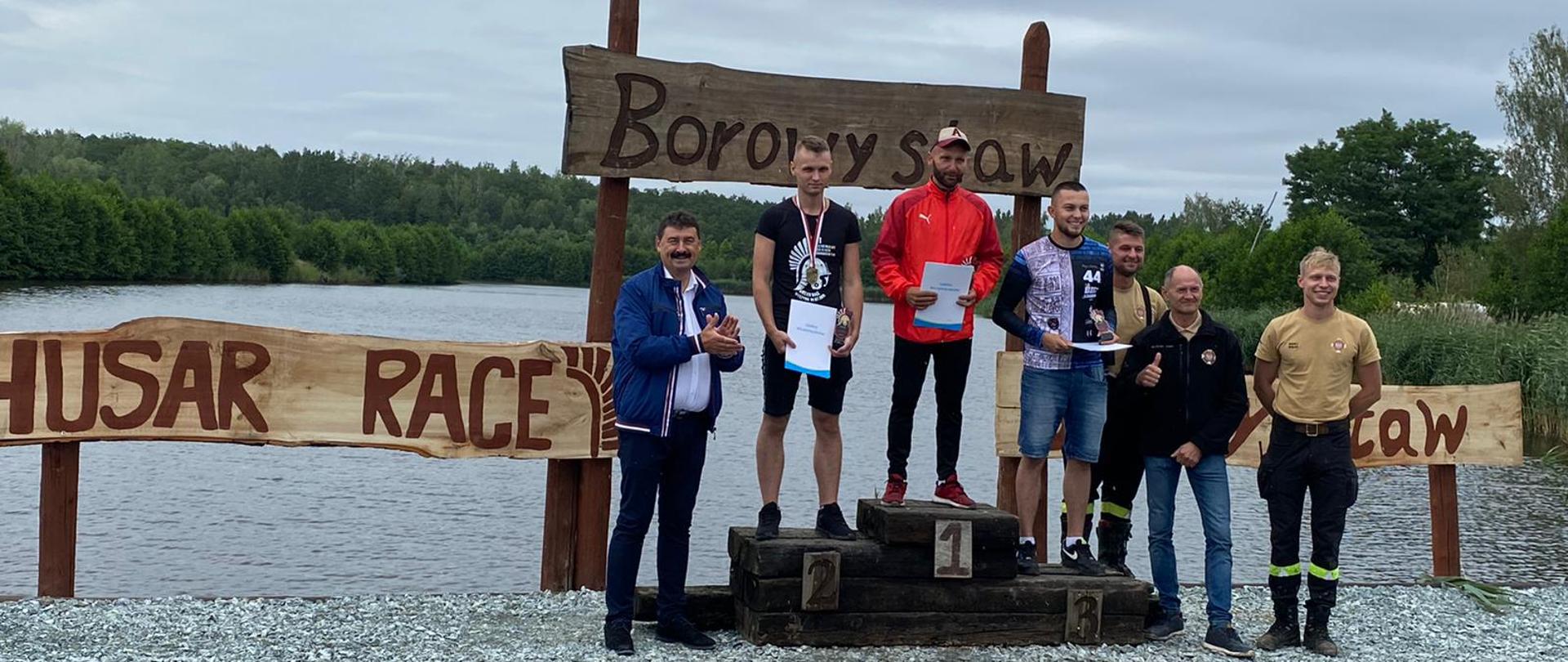 Na zdjęciu podium na nim zawodnicy obok organizatorzy z Sekretarzem Stanu Ministerstwa Rolnictwa i Rozwoju Wsi za nimi jezioro obok podium tablice z napisem Husar Race a za podium napis Borowy Staw